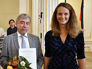 Juri Rosov und Bettina Martin stehen nebeneinander. Juri Rosov hält Blumen und eine Urkunde in der Hand.