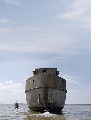 Ein altes Schiff liegt mit dem Heck nach vorn im seichten Wasser. Neben dem Boot geht ein Mann durchs Wasser.