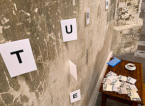An der Wand hängen Papierblätter. Jedes enthält einen Großbuchstaben. Vor der Wand steht ein brauner Tisch. Darauf befinden sich eine Kaffeetasse, eine Medikamentenbox und viele Zettel mit der gedruckten Aufschrift „Nicht vergessen“ und handschriftlichen Notizen.  