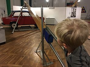 Ein Kind drückt einen überdimensionalen Hebelarm nach unten. Am Ende des Hebels schwebt ein roter Trabant.