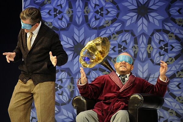 Ein Schauspieler sitzt mit einer Schlafmaske über den Augen in einem Sessel. Hinter ihm befindet sich eine Tuba. Neben ihm tanzt ein Mann. Auch er trägt eine Schlafmaske.
