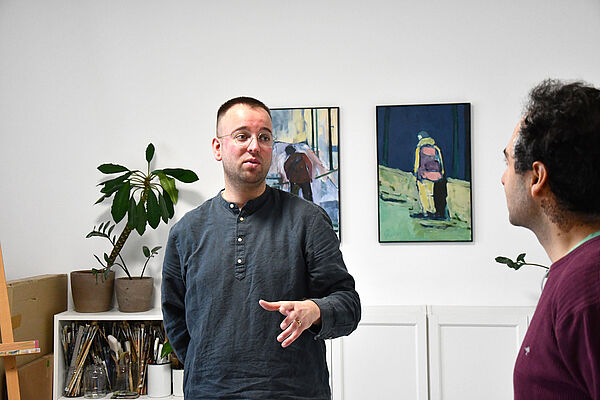 Das Foto zeigt Felix Jes vor 2 Malereien. Im Vordergrund steht ein Mann. Sie unterhalten sich.