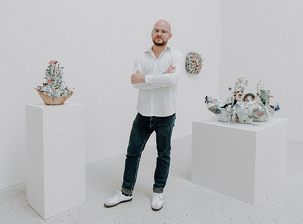 Ein Mann steht lässig zwischen mehreren Skulpturen in einem weißen Ausstellungsraum.