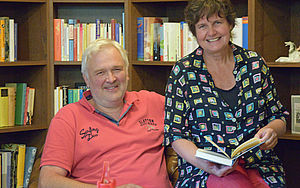 Torsten und Conny Brock sitzen vor einem Bücherregal. Conny Brock hält ein aufgeschlagenes Buch in der Hand. 