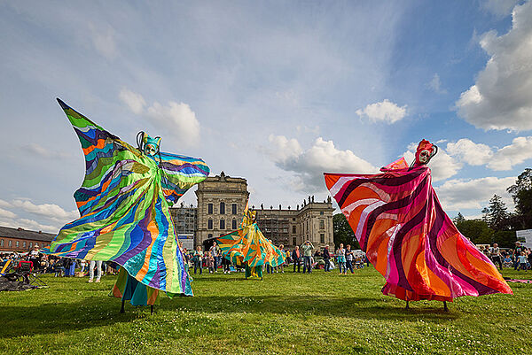 Zwei Künstler auf Stelzen. Sie tragen bunte Kleider. Sie tanzen tanzen auf einer Wiese. Im Hintergrund steht das Schloss von Ludwigslust.