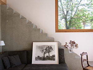 Ein Raum. Mit einem Ausschnitt eines quadratischen Fensters. Davor führt eine graue Treppe einen Stock höher. Vor der Treppe steht ein braunes Sofa. Darauf steht ein Bilderrahmen mit einer Schwarz-Weiß-Fotografie zweier Bäume.