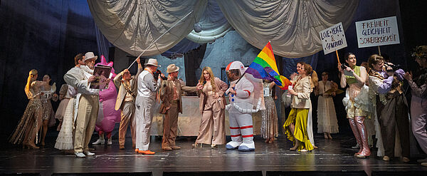 Bühnenszene mit vielen Menschen. Im Zentrum eine Frau in rosa Hosenanzug neben einer aufblasbaren Astronautenfigur mit Regenbogenfahne. Rechts im Bild Schilder mit den Aufschriften „Love Peace & Gravitation" und „Freiheit Gleichheit Schwerelosigkeit".