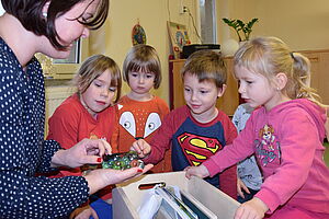 Vier Kinder hocken um eine Holzkiste. Johanna Bojarra hält ein Netz voller Murmeln in der Hand und zeigt es den Kindern.