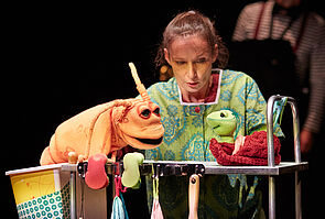 Eine Frau steht an einem kleinen Metalltisch. Mit ihren Händen führt sie Handpuppen.