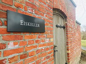 Nahaufnahme einer Backsteinwand mit einer Holztür. Neben der Tür hängt ein Schild mit der Aufschrift "Eiskeller".