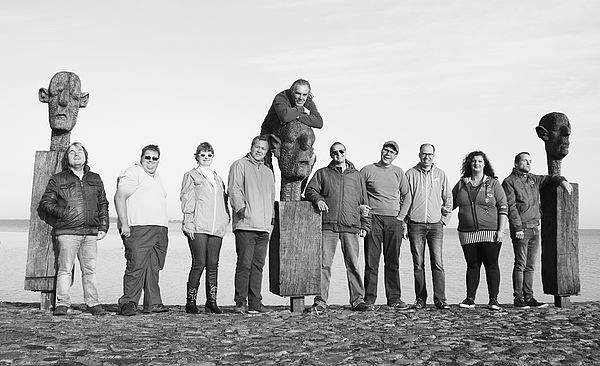 Die zehn Musikerinnen und Musiker von „Seeside“ stehen neben drei großen Holzskulpturen am Strand. 