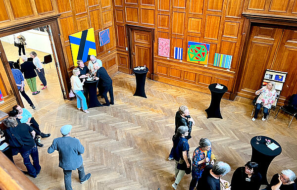 Blick von der Galerie in einem wandgetäfelten Ausstellungsraum. Menschen stehen an Tischen, erzählen oder laufen herum. An der Wand hängen Kunstarbeiten.