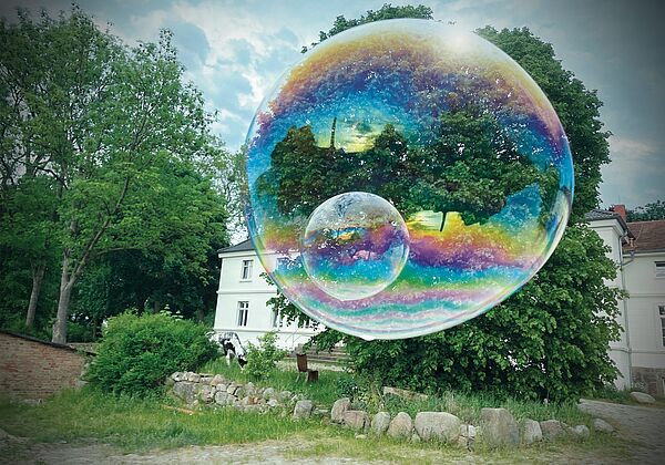 Der weiße Gutshof in Bröllin. Davor stehen Bäume. Davor schwebt eine große Seifenblase in Regenbogenfarben.