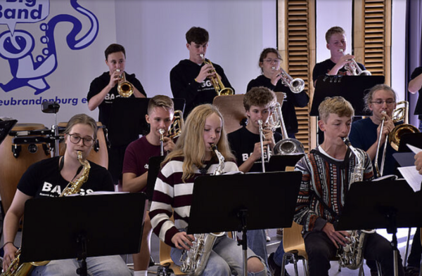 Zehn junge Leute sitzen in einem Raum. Sie haben Instrumente und machen Musik.