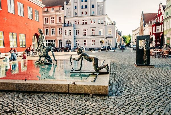 Ein Marktplatz. Im Vordergrund ein Brunnen mit Figuren. Im Hintergrund Häuser.