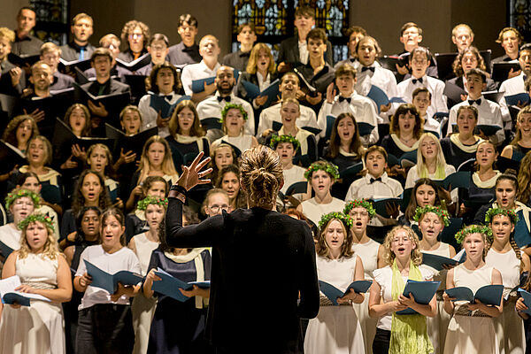 Mehr als 70 Jungen und Mädchen verteilen sich auf acht Reihen. Sie halten Hefter in den Händen und singen. Vor ihnen steht eine Frau. Sie dirigiert.