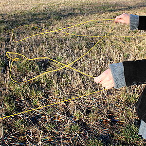 Eine Wiese. Zwei Hände halten einen gelben Faden, der auf der Wiese liegt.