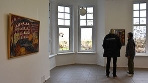 Ein Mann und eine Frau stehen vor großen Fenstern. An der Wand hängt ein Bild.