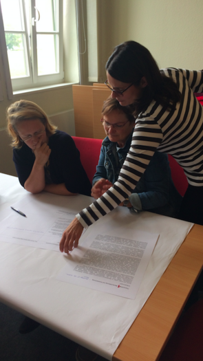 Zwei Frauen sitzen an einem Tisch und lesen einen Zettel. Eine dritte Frau steht neben ihnen und greift nach einem der Zettel, der auf dem Tisch liegt.