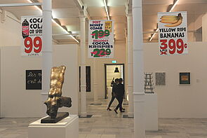 Auf einem Sockel steht eine erotische Skulptur in Gold. Von der Decke hängen Preisschilder für Cola und Bananen.