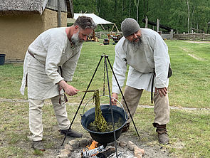 Zwei Männer stehen in historischer Kleidung an einer Feuerstelle. Über dem Feuer hängt ein Topf. 