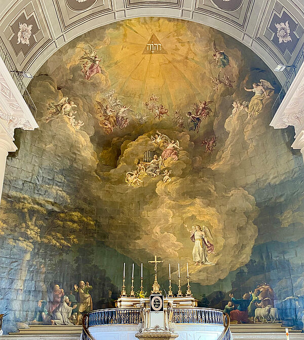 An der Wand befindet sich ein großes Gemälde. Es reicht vom Boden bis zur Decke und bedeckt von links nach rechts die gesamte Wand. Auf dem Gemälde sind Wolken, Engel, Hirten und Schafe.