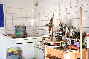 Pinseln und Farben stehen auf einem Tisch. Dahinter ist ein Waschbecken an der Wand.