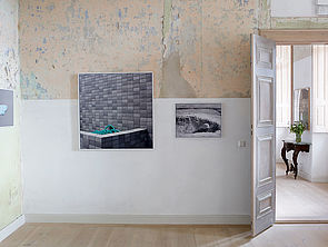 Eine Wand. Einige Stellen sind weiß gestrichen. An anderen Stellen platzt alte Farbe ab. Eine Tür ist geöffnet. An der Wand hängen zwei Fotografien.