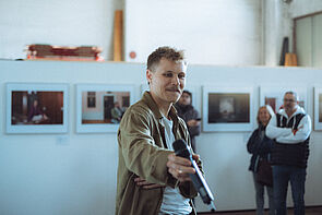 Ein Mann hält ein Mikrofon. Im Hintergrund: eine Bildergalerie.