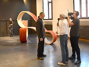 Künstler Broder Burow wird im Ausstellungsraum gefilmt und befragt.