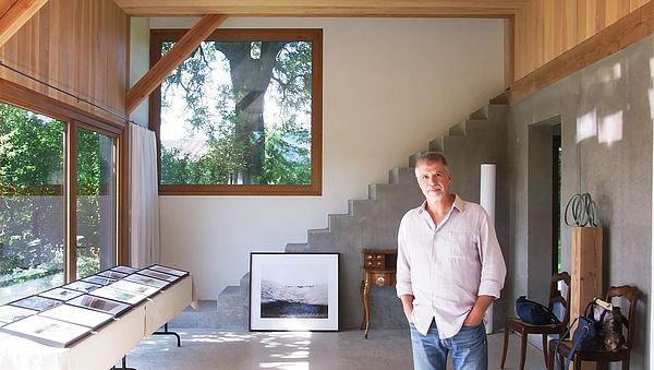 Hans-Christian Schink steht in seinem Atelier. Links befindet sich eine große Glasfront, geradeaus ein quadratisches Fenster. Vor der Glasfront liegen Bilderrahmen auf Tischen. Im Raum, an einer Treppe, lehnt eine große Fotografie.