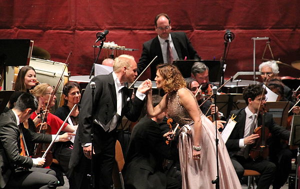 Matthias Koziorowski und Stephanie Elliott halten sich an der Hand, schauen sich an und singen. Im Hintergrund: die Staatskapelle.