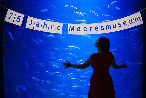 Eine Frau steht vor einem großen Aquarium voller Fische. Auf dem Aquarium hängt eine Girlande mit der Aufschrift 75 Jahre Meeresmuseum