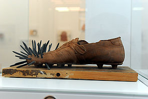 Auf einem Holzbrett steht ein Schuh. Im vorderen Teil des Schuhs stecken Nägel.