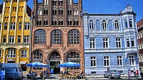 Ein gelbes und backsteinfarbenes Giebelhaus. Daneben ein blaues Eckhaus. Davor: der Marktplatz von Stralsund. Menschen sitzen unter blauen Sonnenschirmen an Restauranttischen. 