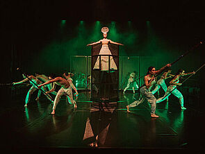 Eine Bühne. Sie ist dunkel und grün angeleuchtet. In der Mitte steht eine Frau. Daneben stehen im Halbkreis Frauen und Männer.