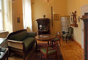 In einer Stube stehen ein historisches Sofa, zwei Sessel und ein runder Tisch. In der Ecke befindet sich ein Kachelofen. An der Wand steht ein dunkler, geöffneter Sekretär.