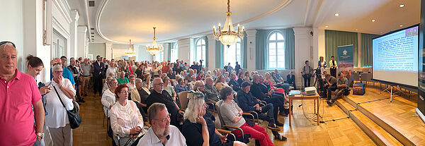 Viele Menschen sitzen in einem großen Saal auf Stuhlreihen und schauen auf eine Bühne am rechten Bildrand, auf der die Übertragung der Unesco-Sitzung auf einer Leinwand läuft.