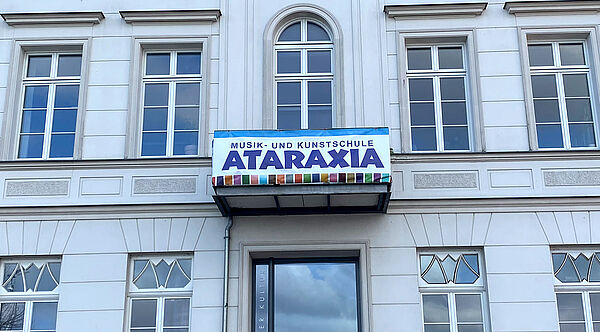 Die Außenfassade von Ataraxia. In der Mitte hängt ein Balkon, daran ein Banner mit der Aufschrift "Ataraxia".