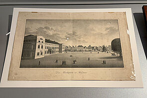 Ein großer Platz. Auf dem Platz sind Menschen. Links und im Hintergrund stehen Häuser. Rechts steht ein Baum. 