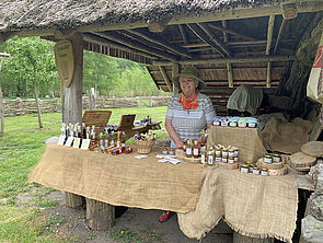 Eine Frau steht hinter einem Holztisch voller Flaschen und Gläser und verkauft Öle, Marmeladen und Tinkturen.