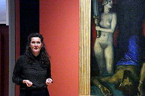 Eine Schauspielerin steht vor einem Gemälde und spricht zum Publikum.