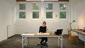 Ein Raum mit wandbreiten Fenstern. In der Mitte steht ein Tisch. Daran sitzt Susanne Gabler und arbeitet.