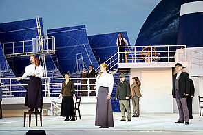 Zehn Schauspielerinnen und Schauspieler stehen an Deck und singen. 