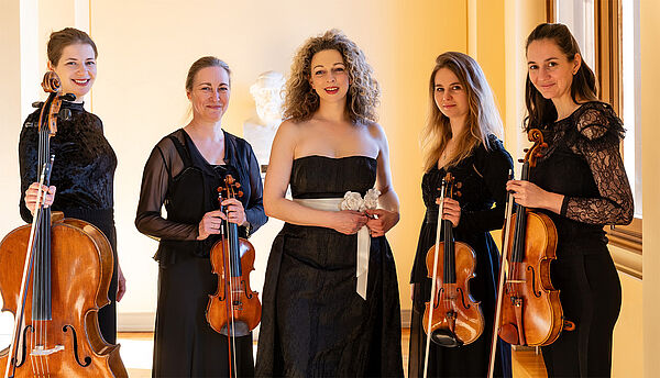 Fünf Frauen stehen nebeneinander. Vier Frauen haben ein Instrument. Die Frau in der Mitte nicht. Sie ist die Sängerin.