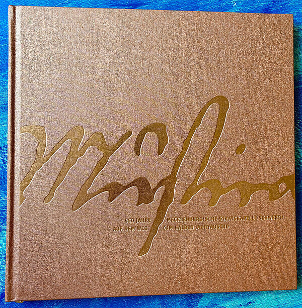 Ein bronzefarbenes Buch-Cover. Auf dem Cover steht: 450 Jahre Mecklenburgische Staatskapelle – Auf dem Weg zum halben Jahrtausend.