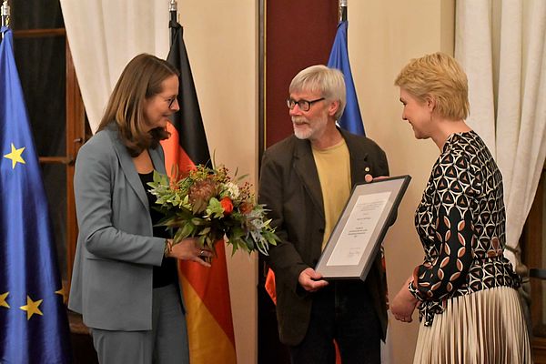 Wolf Karge steht mit der Urkunde in der Hand zwischen Kulturministerin Bettina Martin und Ministerpräsidentin Manuela Schwesig. Bettina Martin hält einen Blumenstrauß in der Hand.