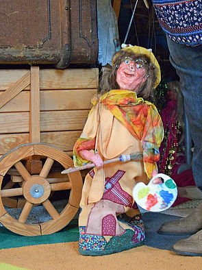 Eine Marionette, die eine Malerin darstellt. In ihren Händen hält sie eine Mischpalette und einen Pinsel.