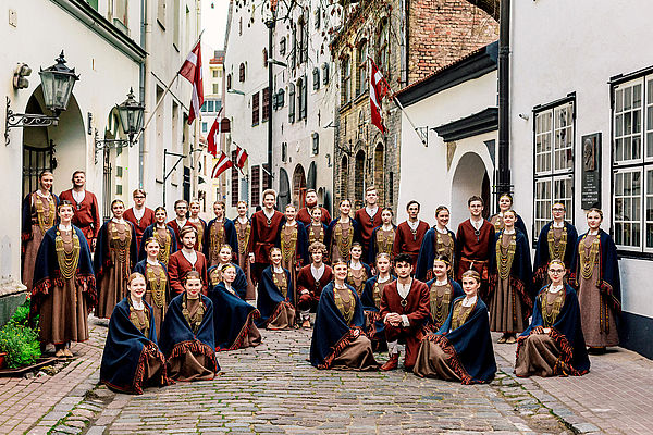 Etwa 40 Sängerinnen und Sänger stehen oder hocken in traditionellen Trachten in einem Halbkreis in einer lettischen Altstadt-Gasse.