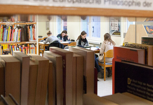 Ein Regal voller Bücher. Dahinter sitzen vier Frauen an Lesetischen.